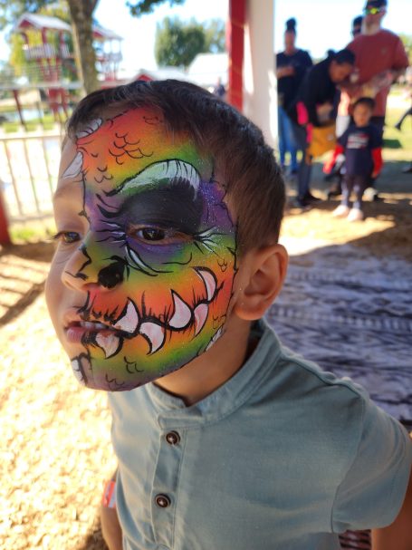 Child with colorful face paint resembling a monster, smiling in a festive environment.