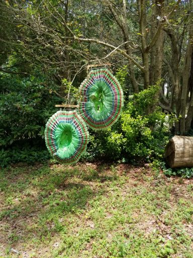 Two colorful woven baskets hanging from a tree branch in a lush green setting.