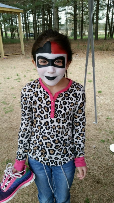 Girl wearing a leopard print shirt and face paint, holding a shoe, standing outdoors.