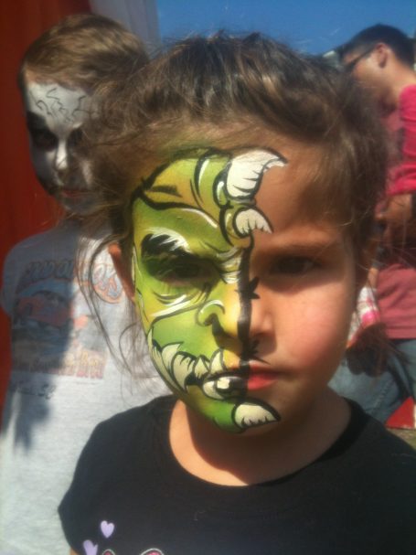 Girl with half her face painted green, resembling a monster, with a serious expression.