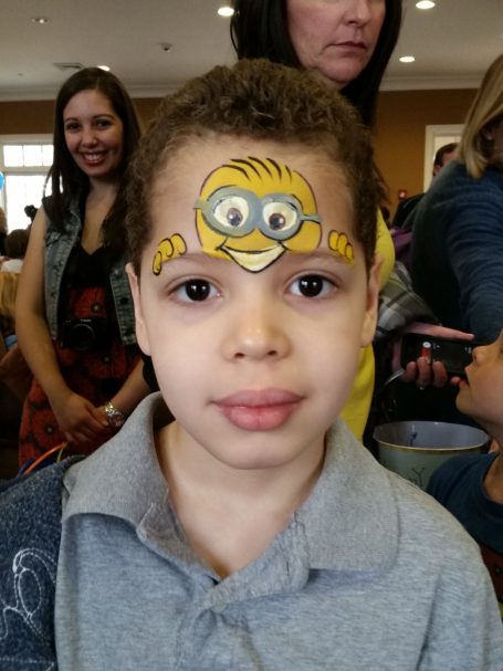 Child with a face painting of a minion on his forehead, smiling in a crowded room.