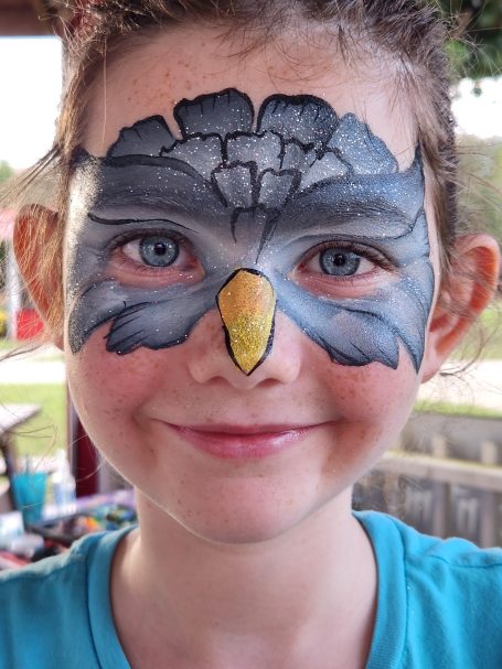 Child with face paint featuring a bird design and bright blue eyes, smiling.