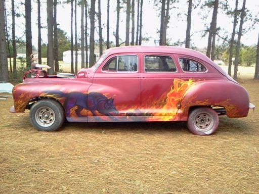 A vintage car with a fiery paint design parked in a wooded area.