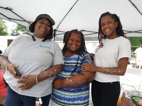 Three smiling individuals stand together under a tent, with face painted arms in script, enjoying a community event.
