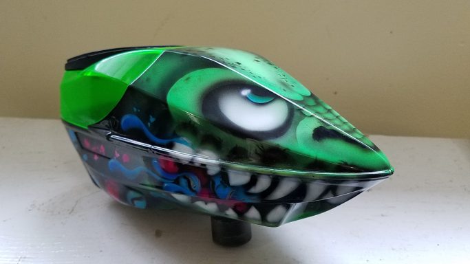A green and black paintball mask featuring monster-themed graffiti.
