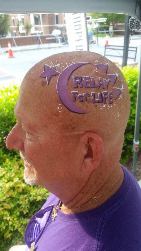 A bald man with a purple "Relay For Life" design on his head, next to a street.