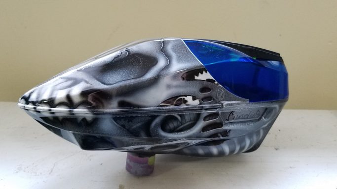 A gray and blue paintball hopper with a skull and swirling designs.