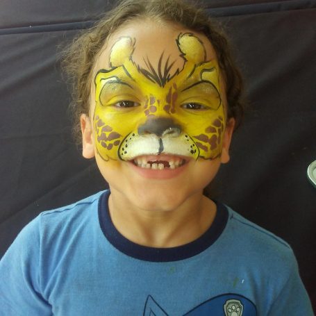 Child with face painted as a giraffe, wearing a blue shirt, smiling broadly.