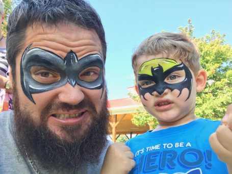 Man and child with Batman face paint, both smiling and posing outdoors.