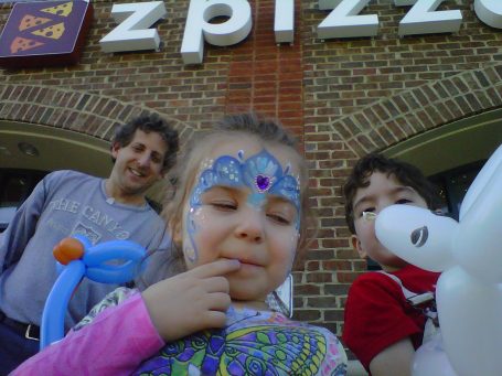 Child with face paint and balloons, with two adults in the background outside a pizza restaurant.