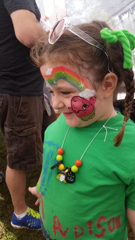 Child with face paint featuring a rainbow and ladybug, wearing a green shirt and colorful necklace.